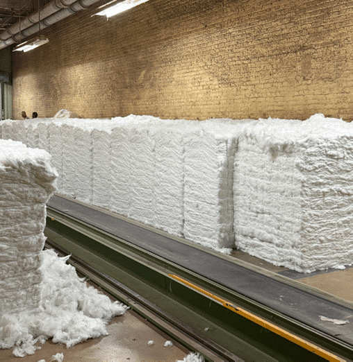 Large stacks of compressed white cotton bales are lined up along a green conveyor belt in a factory with brick walls and overhead lighting. Loose cotton pieces are scattered on the floor.