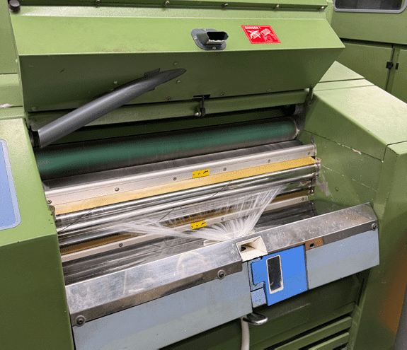 A close-up of a green industrial machine with spinning rollers, tangled white plastic film caught between them, and control buttons visible on the front panel. A warning sticker is displayed above the rollers.